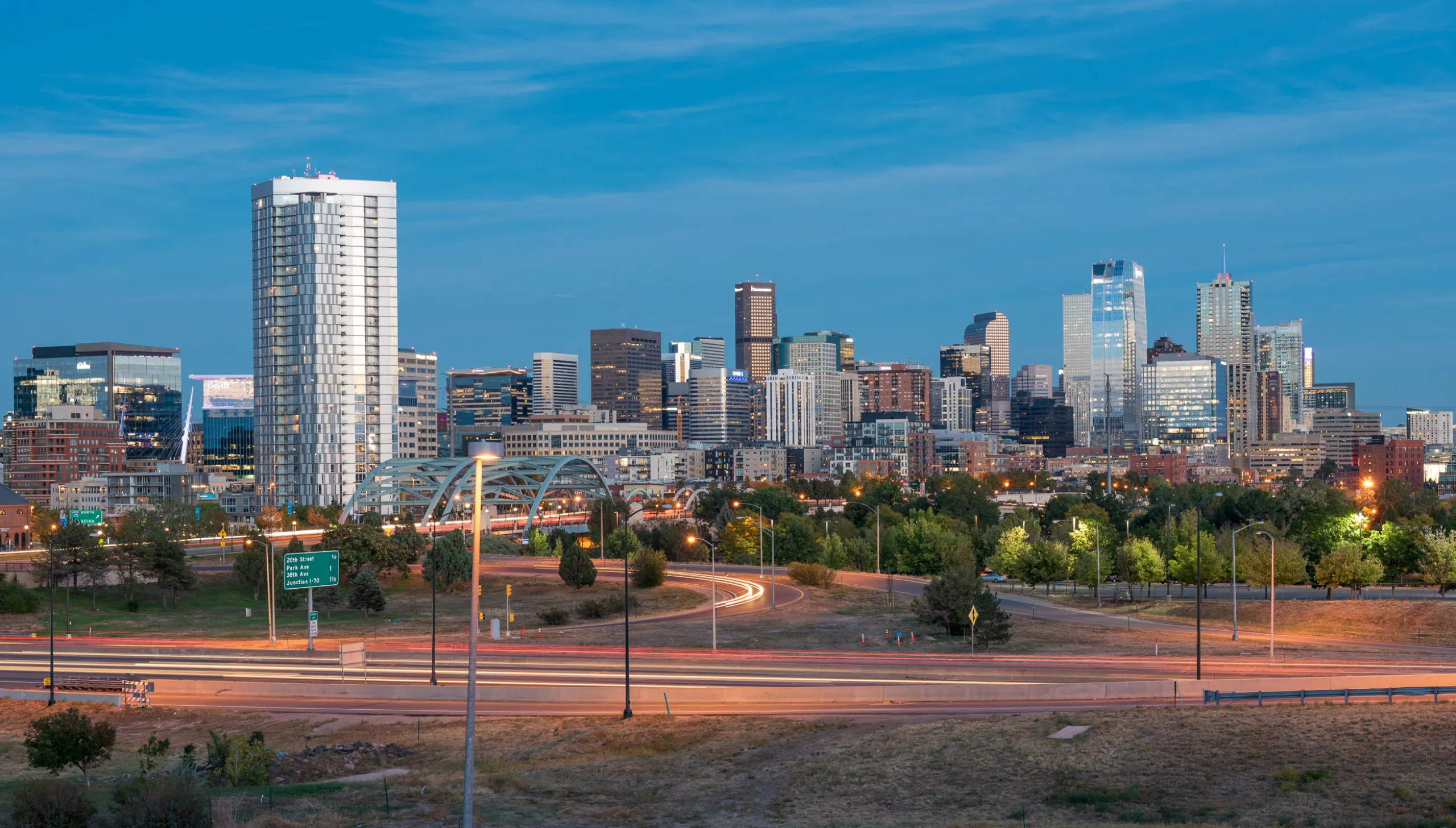 An evening aerial view of the Downtown Denver skyline with streetlights lighting a busy highway streaked with headlights