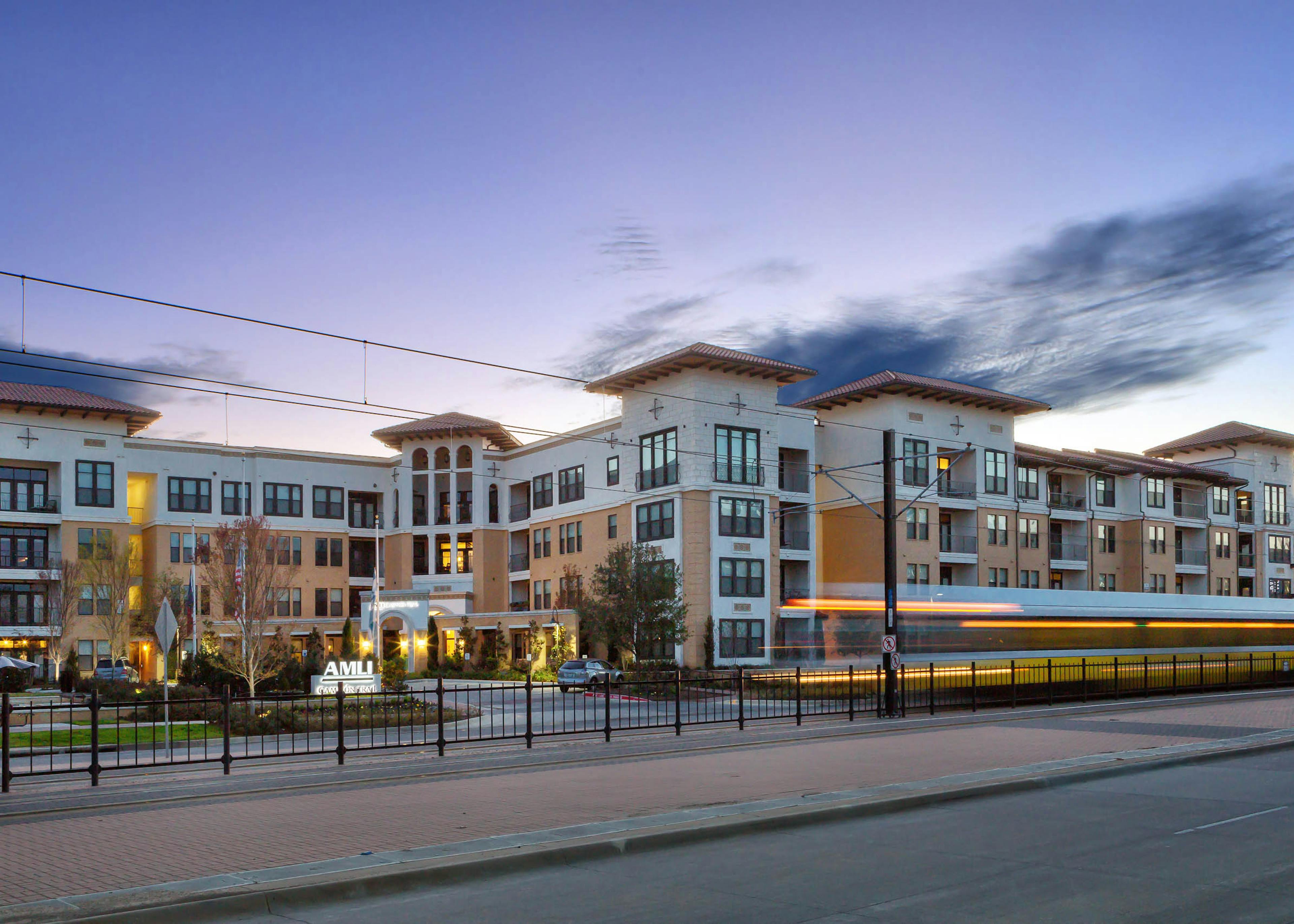 Evening view of blurred DART light rail train along fence traveling in front of the AMLI Campion Trail apartment building