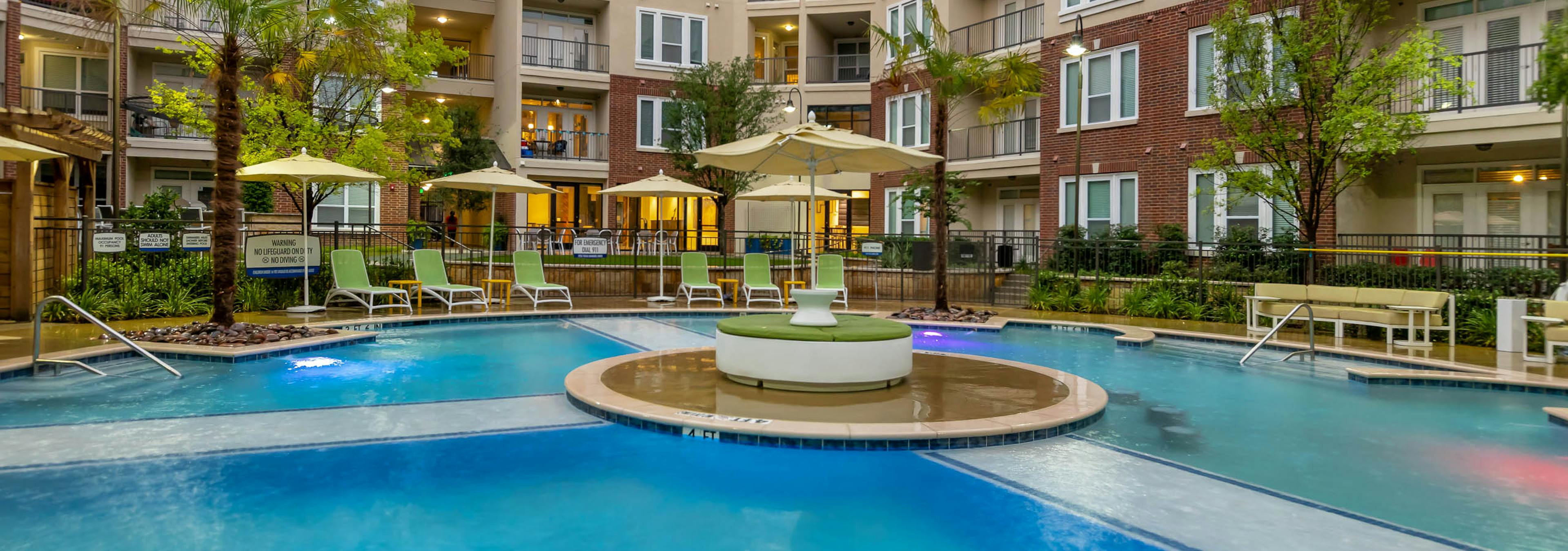 Nighttime view of AMLI Frisco Crossing apartments swimming pool area with lounge seating, umbrellas and palm trees