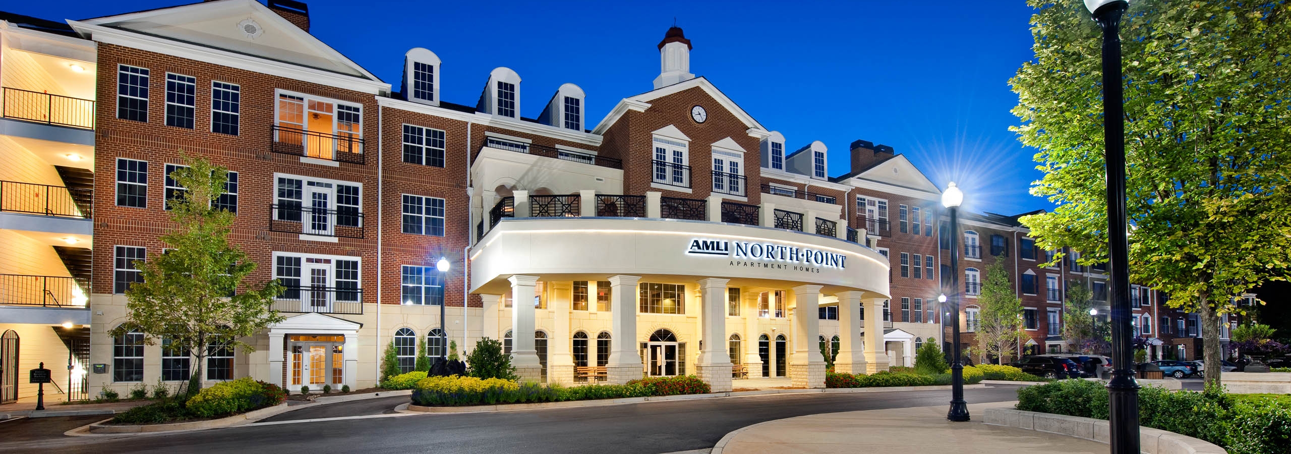 Exterior of AMLI North Point apartment community building at dusk with brick facade and white pillars lining the entry way