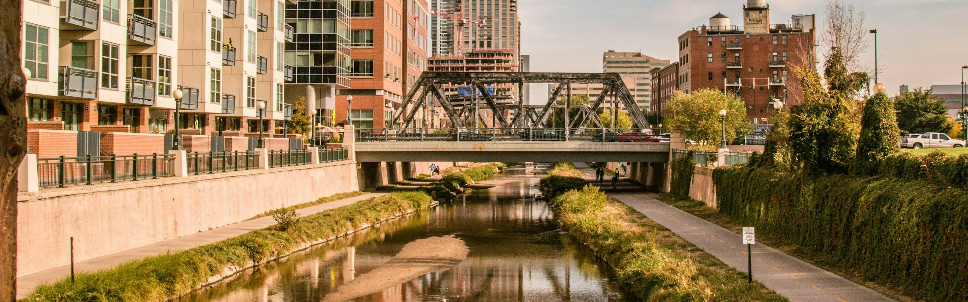 Platte River in downtown Denver view of bike path and pedestrian bridge surrounded by trees and bushes and several buildings 