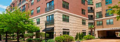 Daytime exterior view of AMLI Uptown brick apartment building and garage entrance and tree lined street with bushes