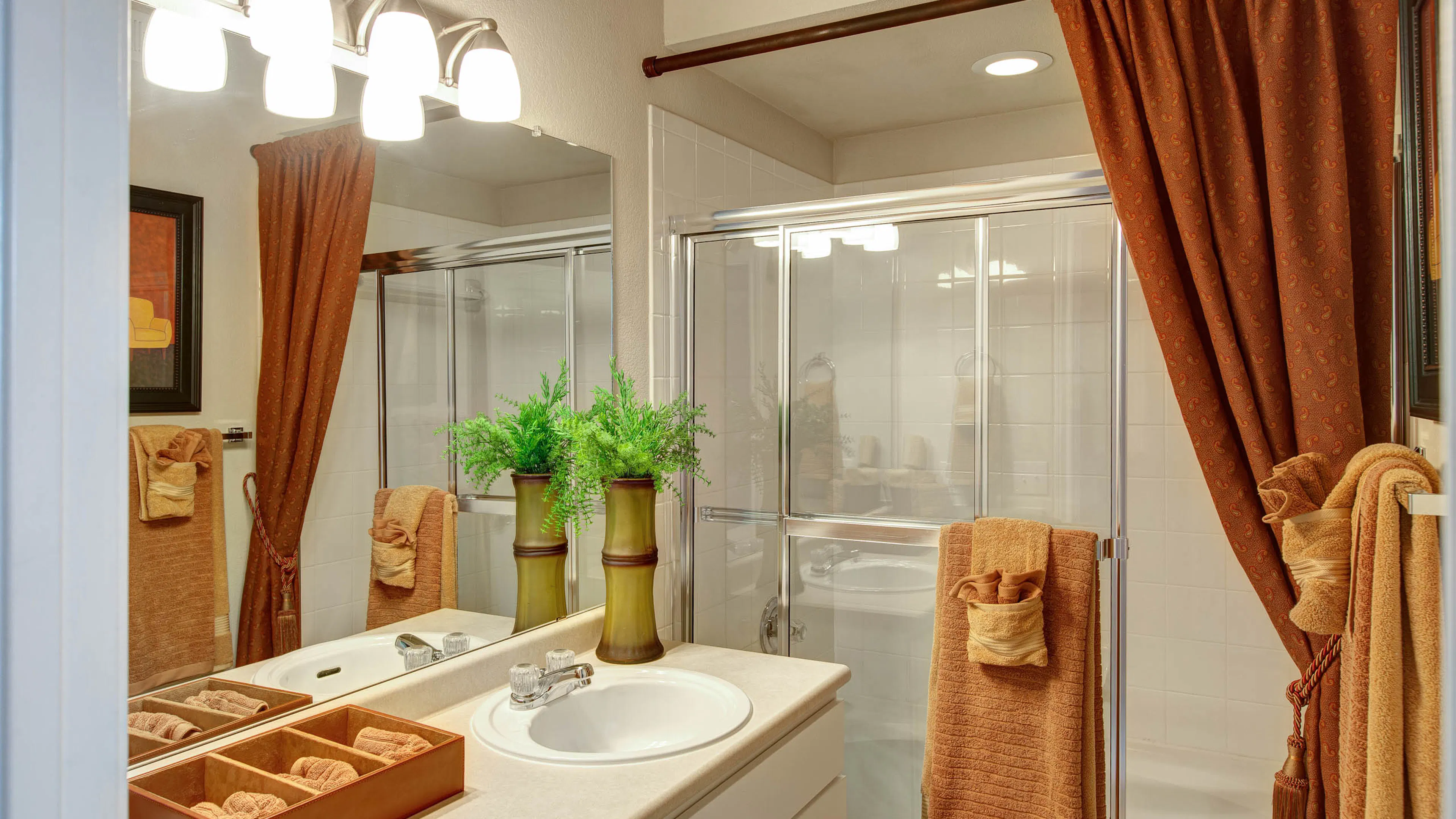 A beige colored bathroom with orange shower curtains and towels and one green plant by the mirror