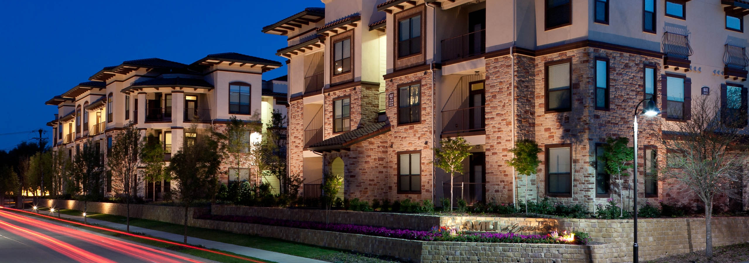 Nighttime exterior view of AMLI at Escena apartment building with lights and trees and red taillights streaks on street