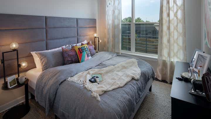 Interior view of AMLI Westside apartment bedroom with queen bed and cream walls with windows and blinds overlooking balcony