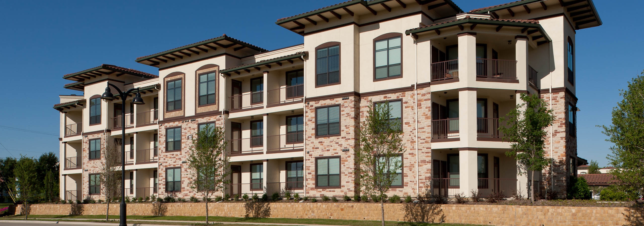Daytime exterior view of AMLI at Escena brick apartment building with low brick wall, tree-lined street and light post