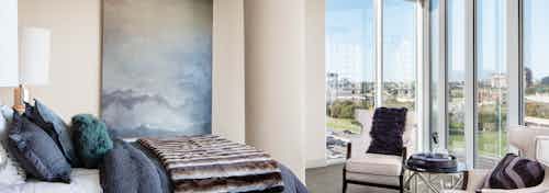 AMLI Design District apartment bedroom with bed and two white chairs and floor to ceiling windows overlooking city