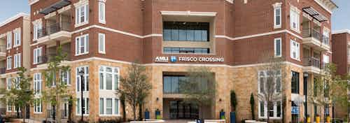 Daytime exterior view of AMLI Frisco Crossing apartment brick facade with sign above building entrance and trees