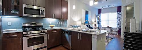 Interior view of AMLI Uptown apartment kitchen with dark brown cabinets and stainless steel appliances and living room