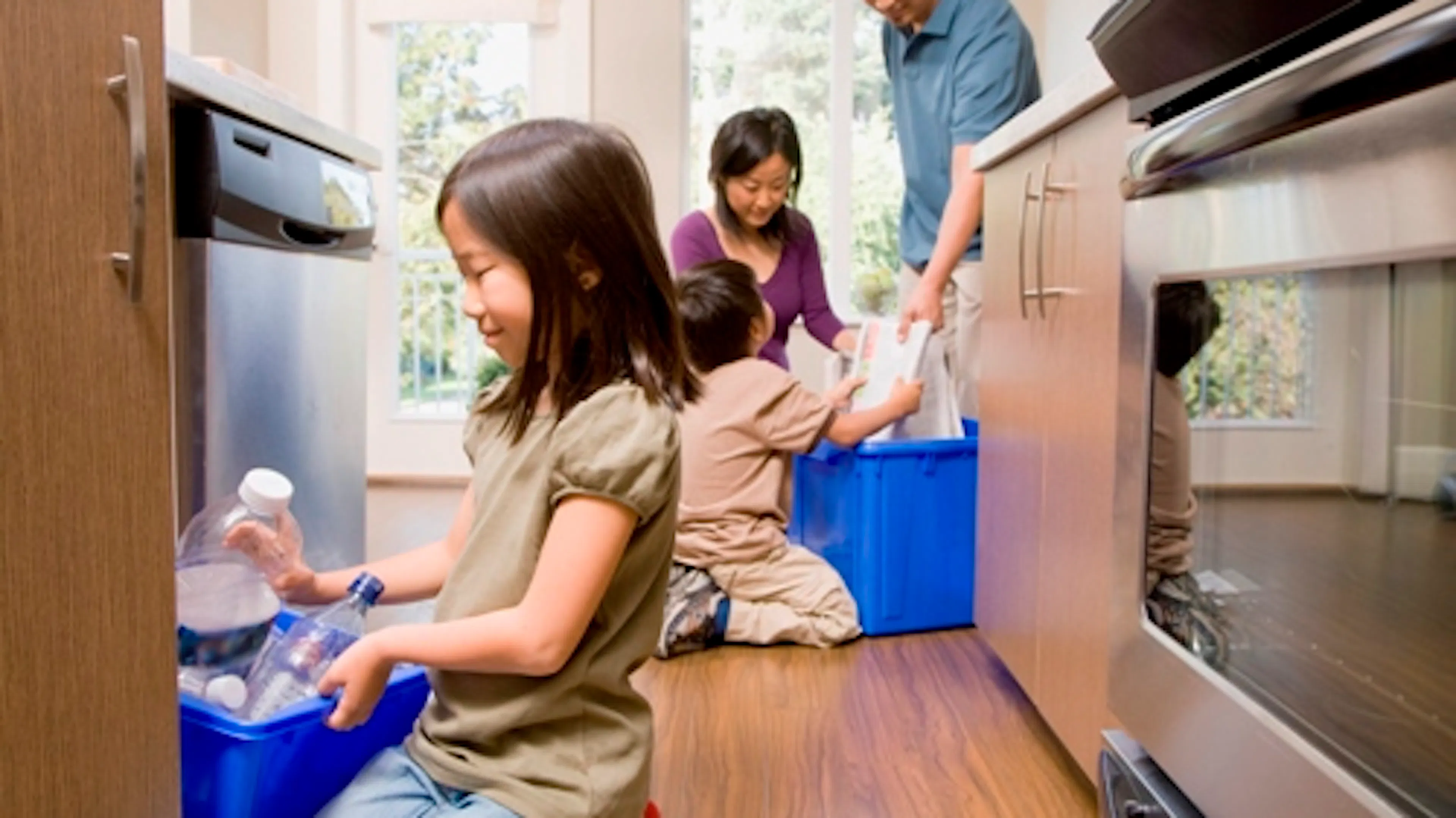 Young girl putting bottles in blue kitchen recycling bin with her family behind her putting other items in a another blue bin