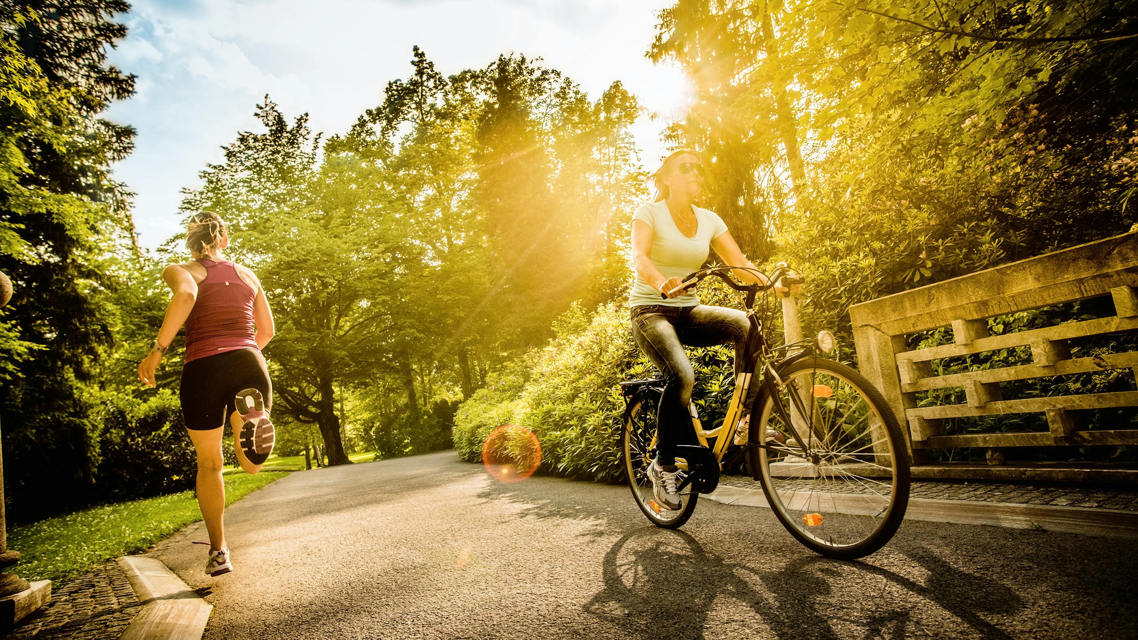 A view of a park with two individuals running and biking down a path with sun shining through lush green trees and bushes