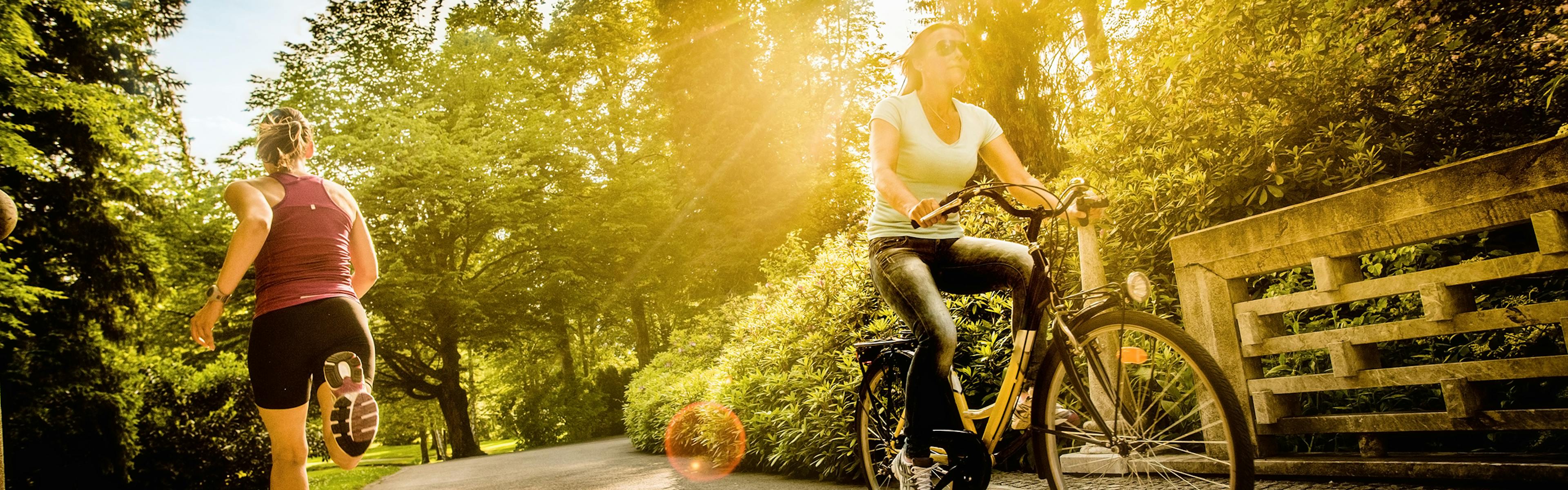 A runner and a biker traveling in opposite directions on a paved park path surround by trees with golden sun shining though