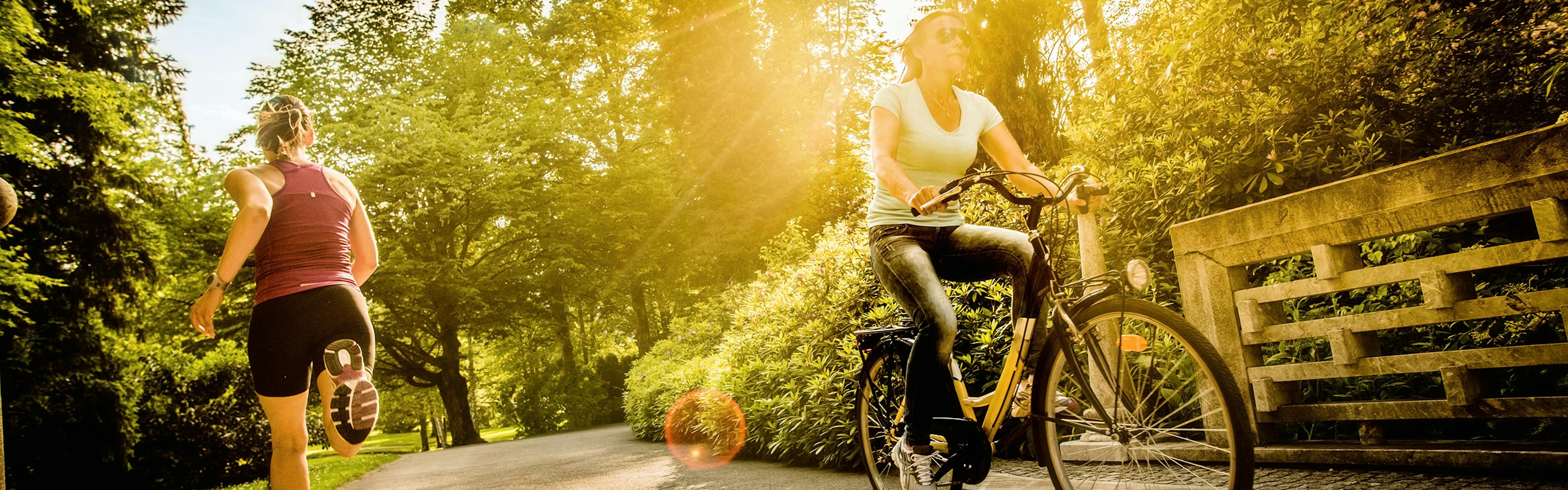 A runner and a biker traveling in opposite directions on a paved park path surround by trees with golden sun shining though