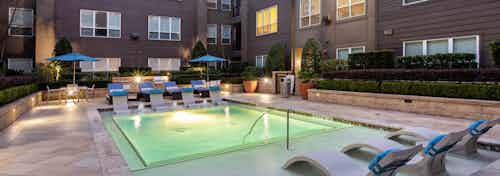Nighttime view of AMLI River Oaks swimming pool with lounge seating and manicured shrubbery encircled by apartment building