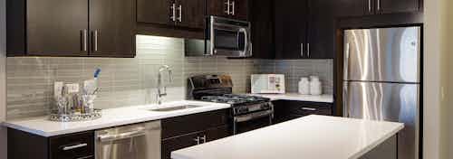AMLI Lofts kitchen with a quartz island alongside dark wooden cabinets and a taupe backsplash with stainless steel appliances