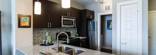 A kitchen at AMLI Arista apartments with a sink and fridge and overhead lighting as well as the front door in the background