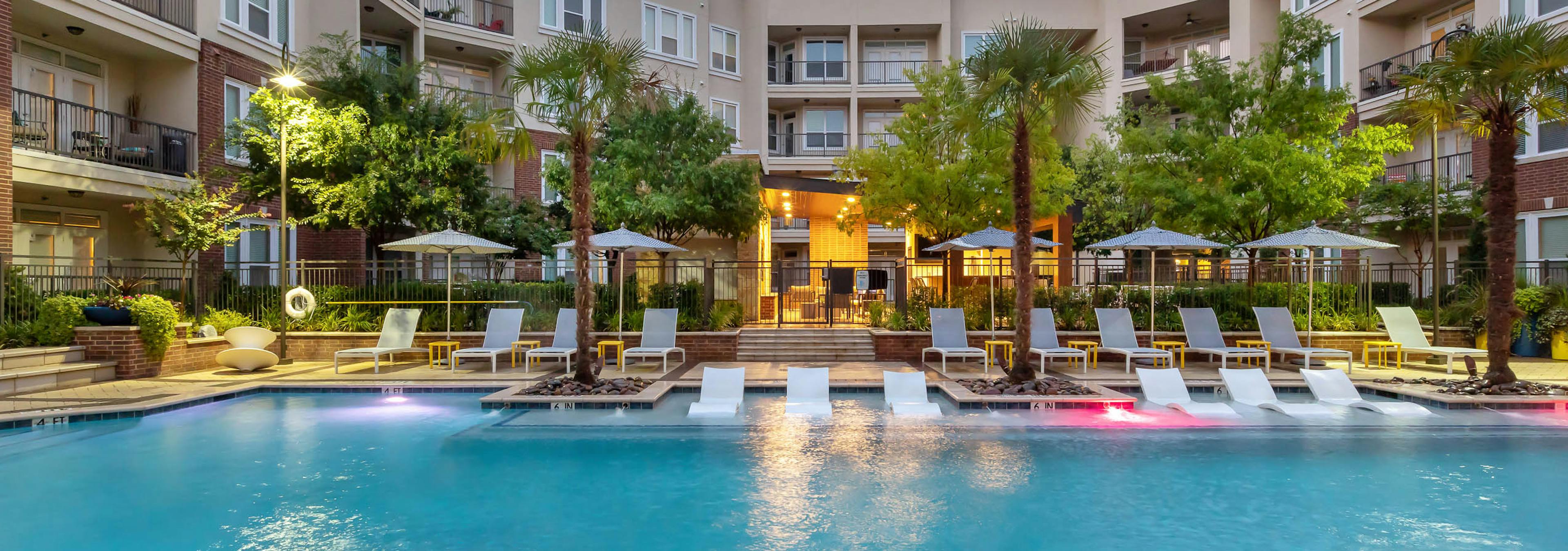 Nighttime view of AMLI Frisco Crossing apartments swimming pool area with lounge seating, umbrellas and palm trees