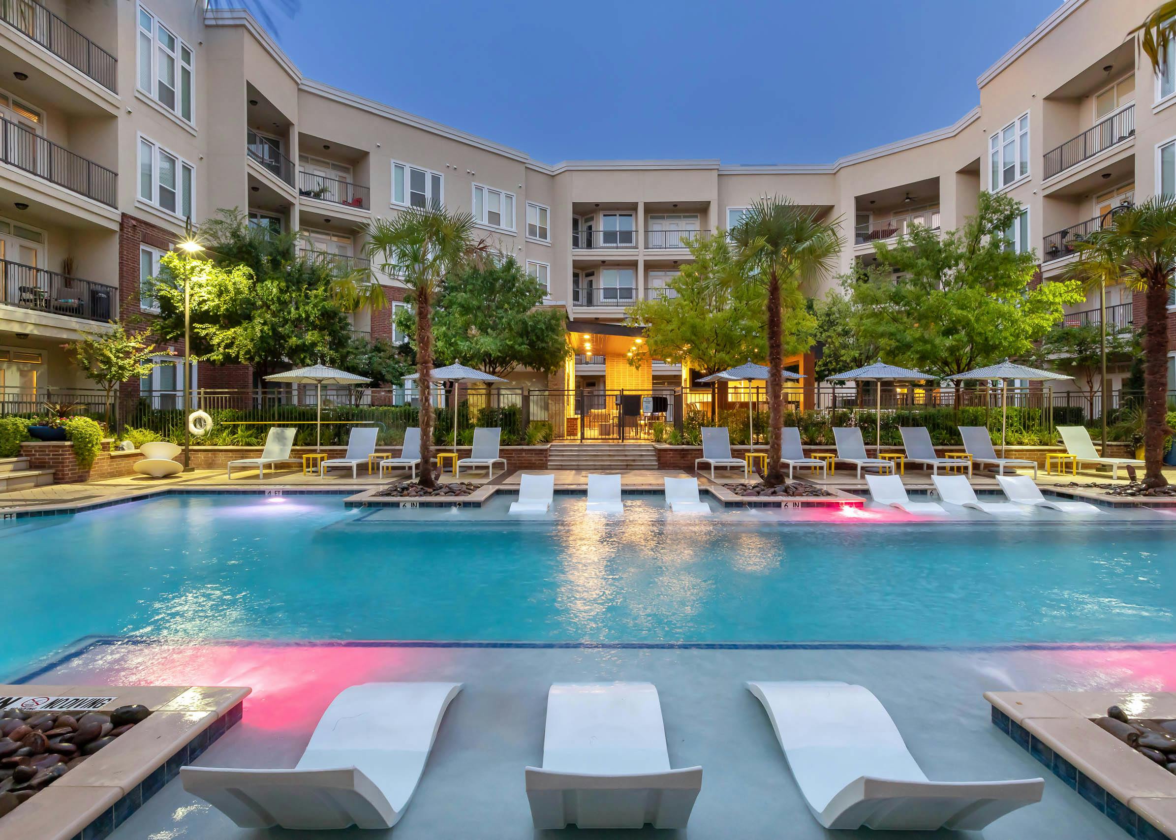 Dusk view of pool area at AMLI Frisco Crossing apartments with lounge seating and umbrellas and lush palm and green trees