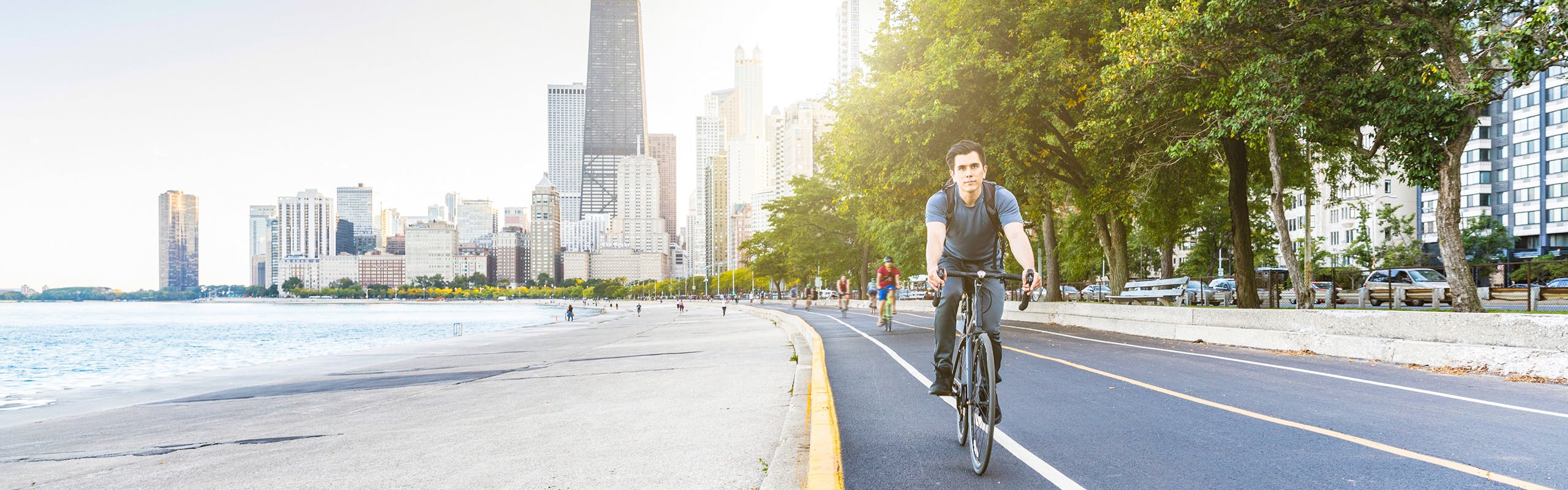Bicycle riders along the pavement beside the lakefront with the sun shining brightly on the Chicago skyline in the background
