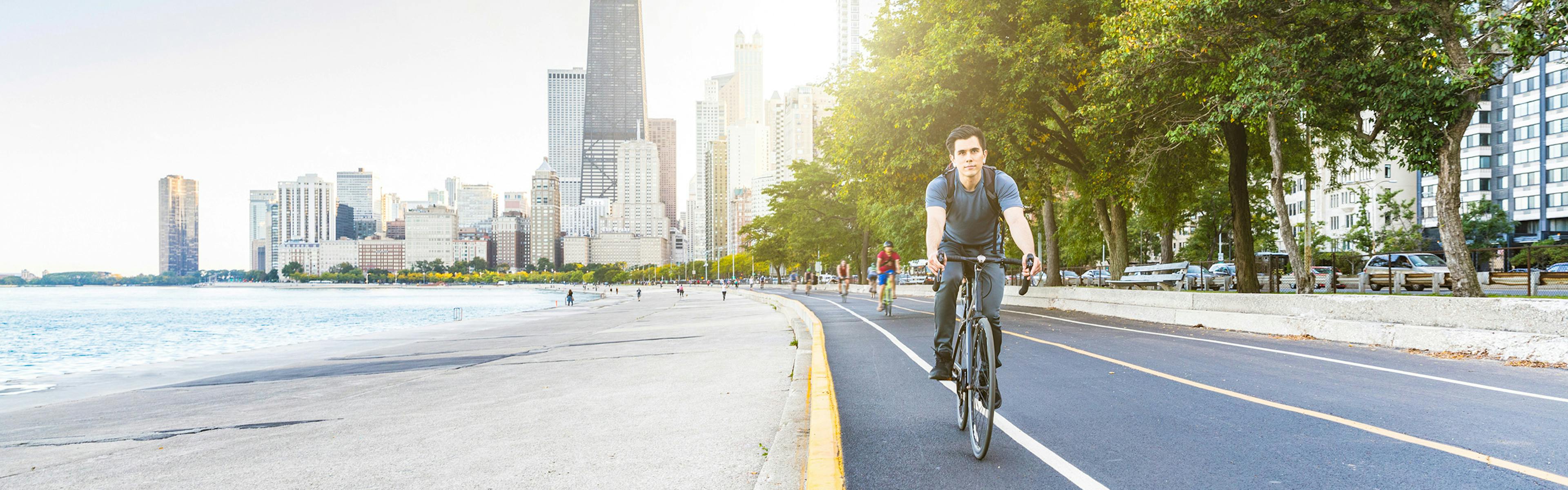 Bicycle riders along the pavement beside the lakefront with the sun shining brightly on the Chicago skyline in the background