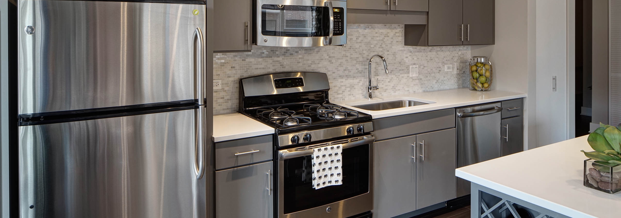An AMLI Lofts kitchen containing stainless steel appliances and grey cabinets paired with quartz countertops and a backsplash