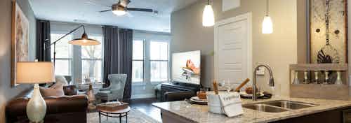 Interior of AMLI Campion Trail apartment kitchen countertop looking into furnished living room with ceiling fan and windows