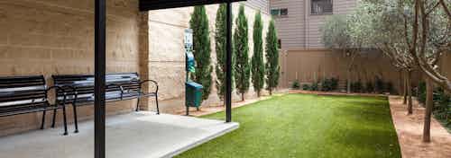 Exterior view of fenced pet park with benches under overhang at left and pet waste station, green grass, trees and shrubbery