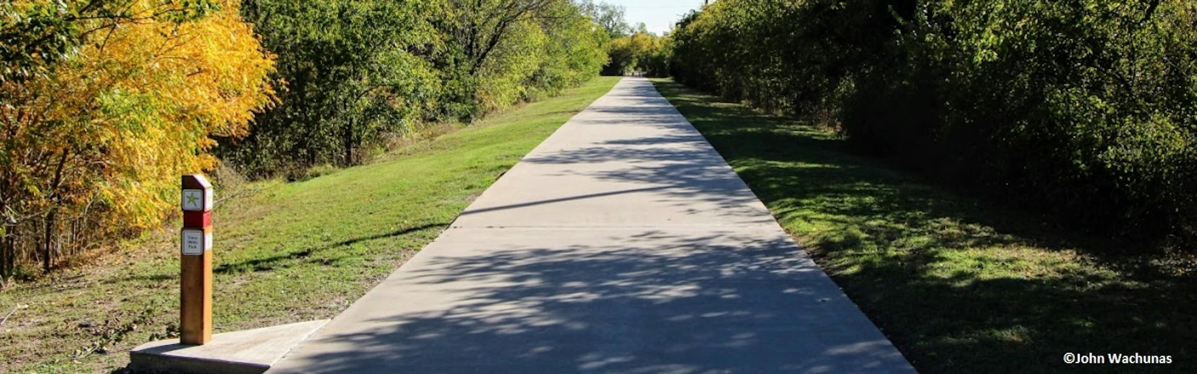 Daytime view of Campion Trail concrete path surrounded on both sides by grass, trees and bushes with sunlight and shadows