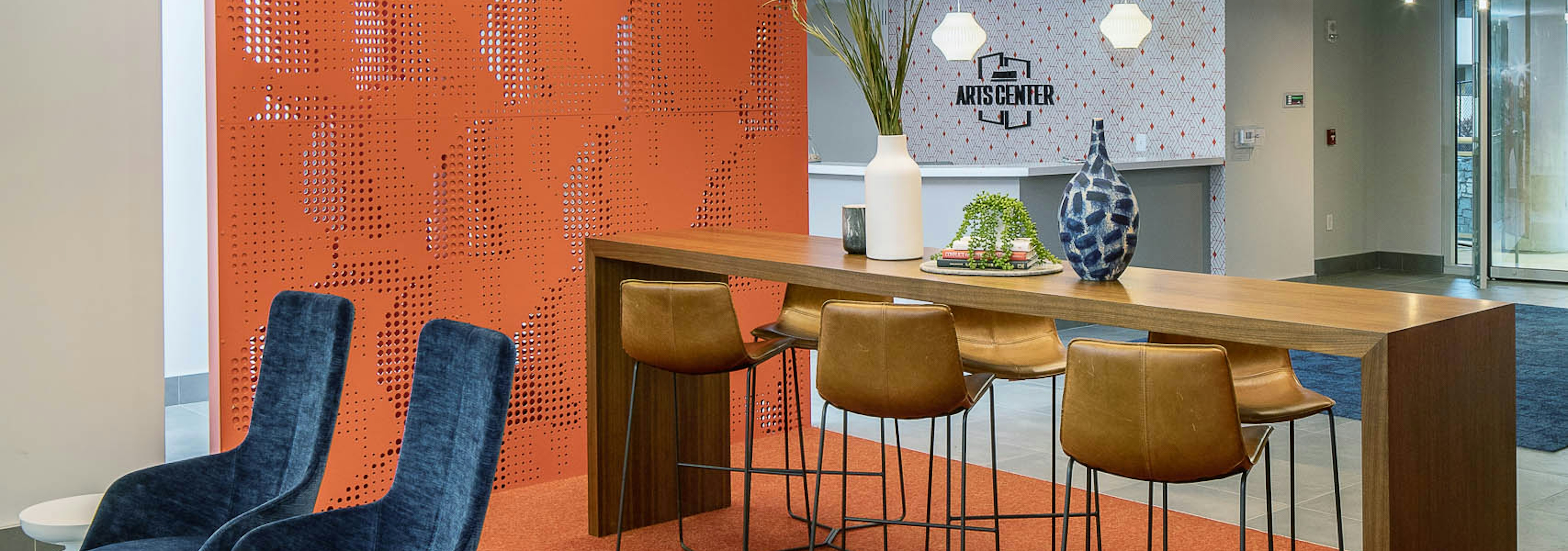 AMLI Arts Center lobby with thin wooden desk and six matching barstools with orange textured wall and modern light fixtures