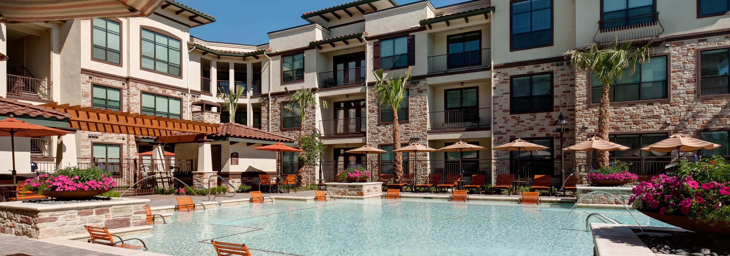 Daytime view of AMLI at Escena apartment building swimming pool with lounge seating in and out of water and umbrellas