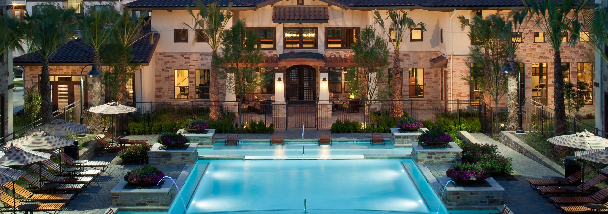 Nighttime view of AMLI at Escena apartments landscaped swimming pool area with lounge seating and umbrellas and palm trees
