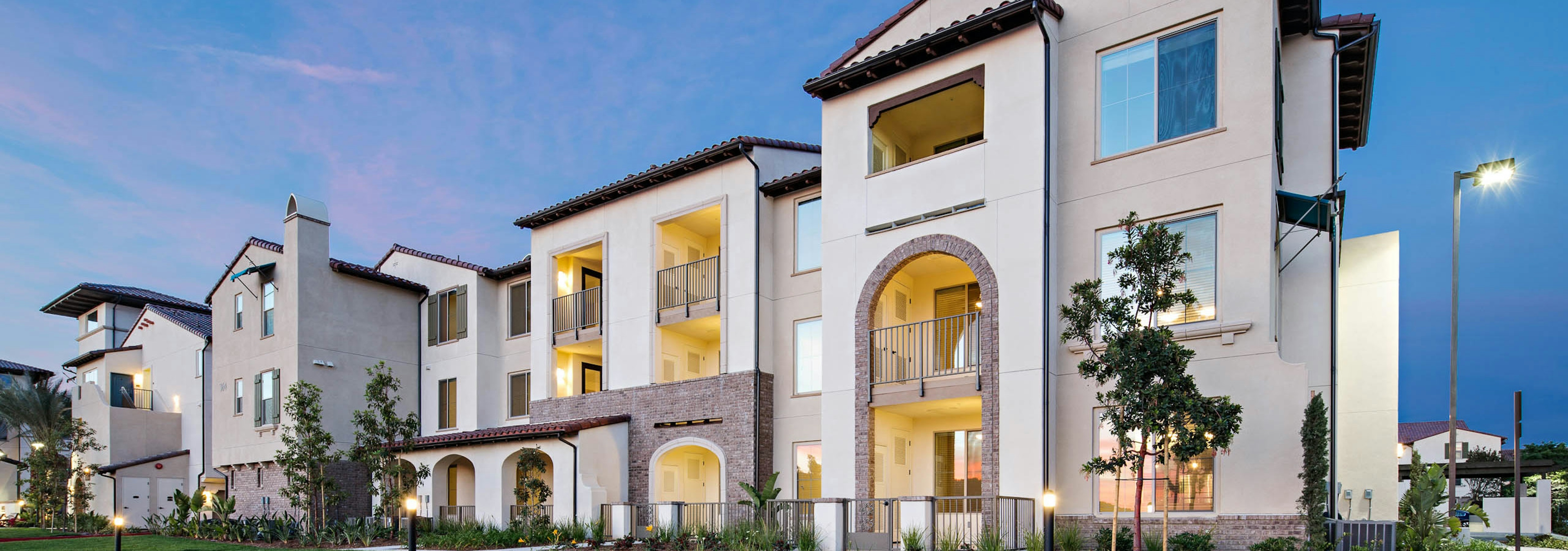 Exterior dusk rendering of AMLI Spanish HIlls apartment building with lit patios and arched entry ways surrounded by greenery