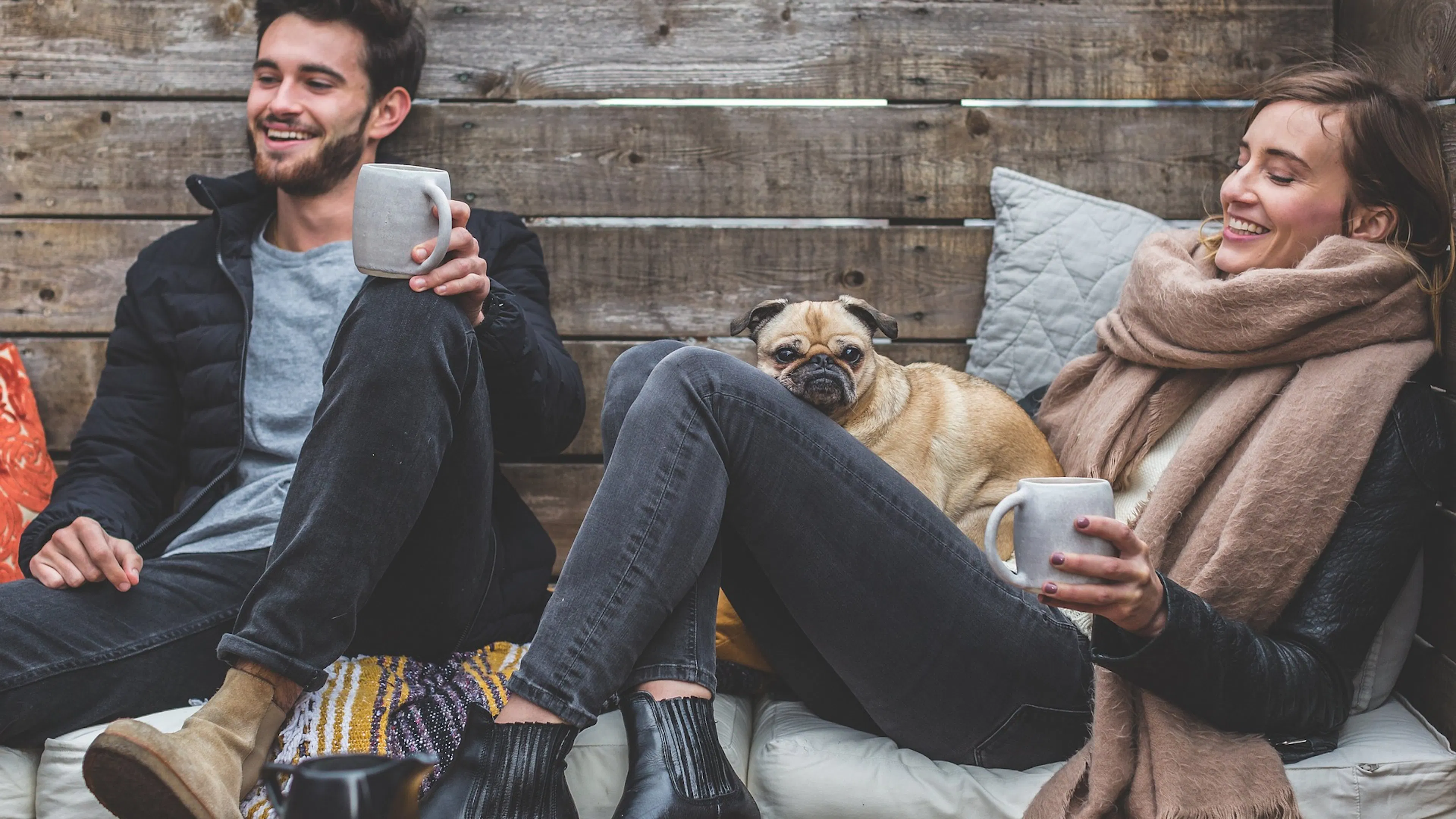 man with coffee cup by woman with scarf and dog sitting by wood wall