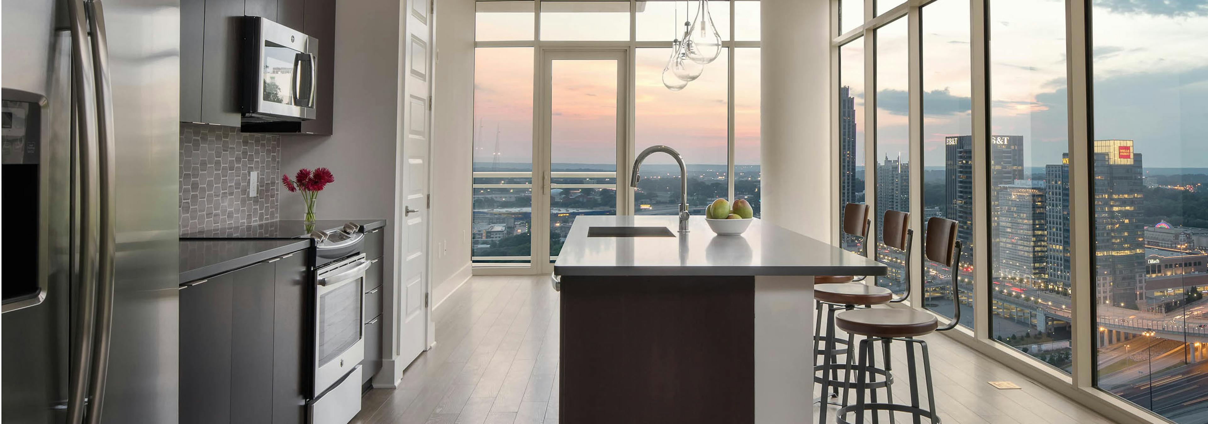 Kitchen at AMLI Arts Center with floor to ceiling windows showing a sunset city view with an island and industrial barstools