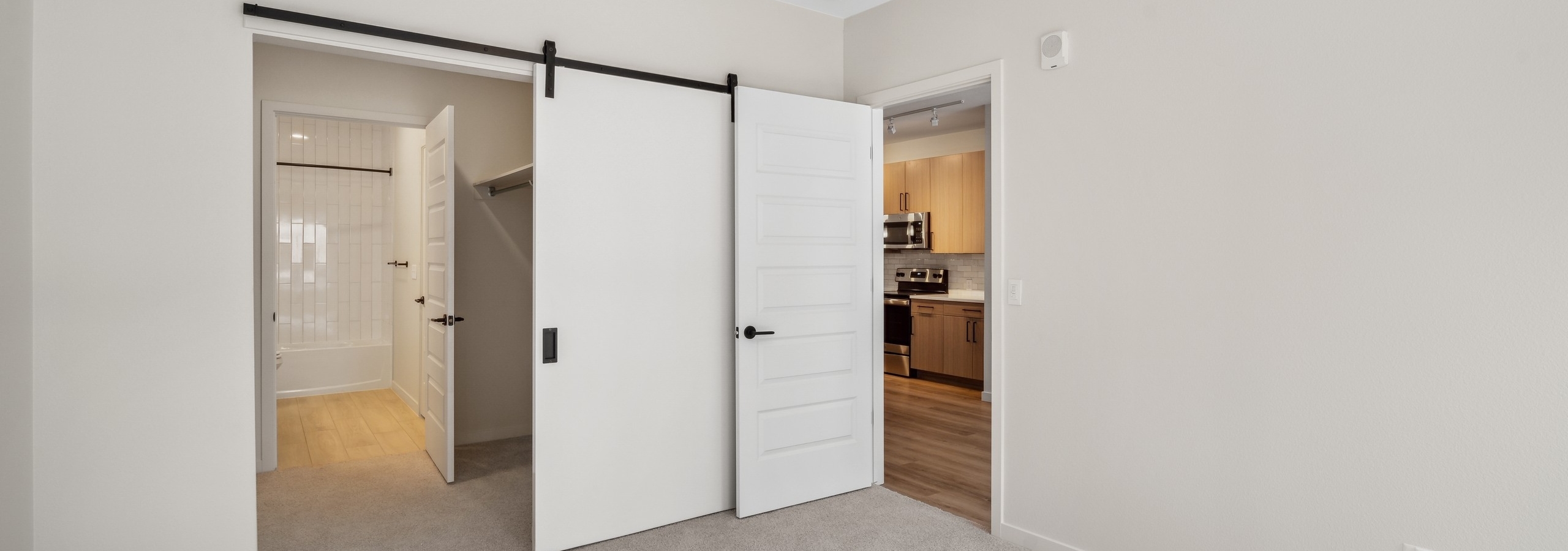 AMLI Broadway Park bedroom with view to kitchen and bathroom with light color carpet and white sliding barn door with black hardware and ceiling fan