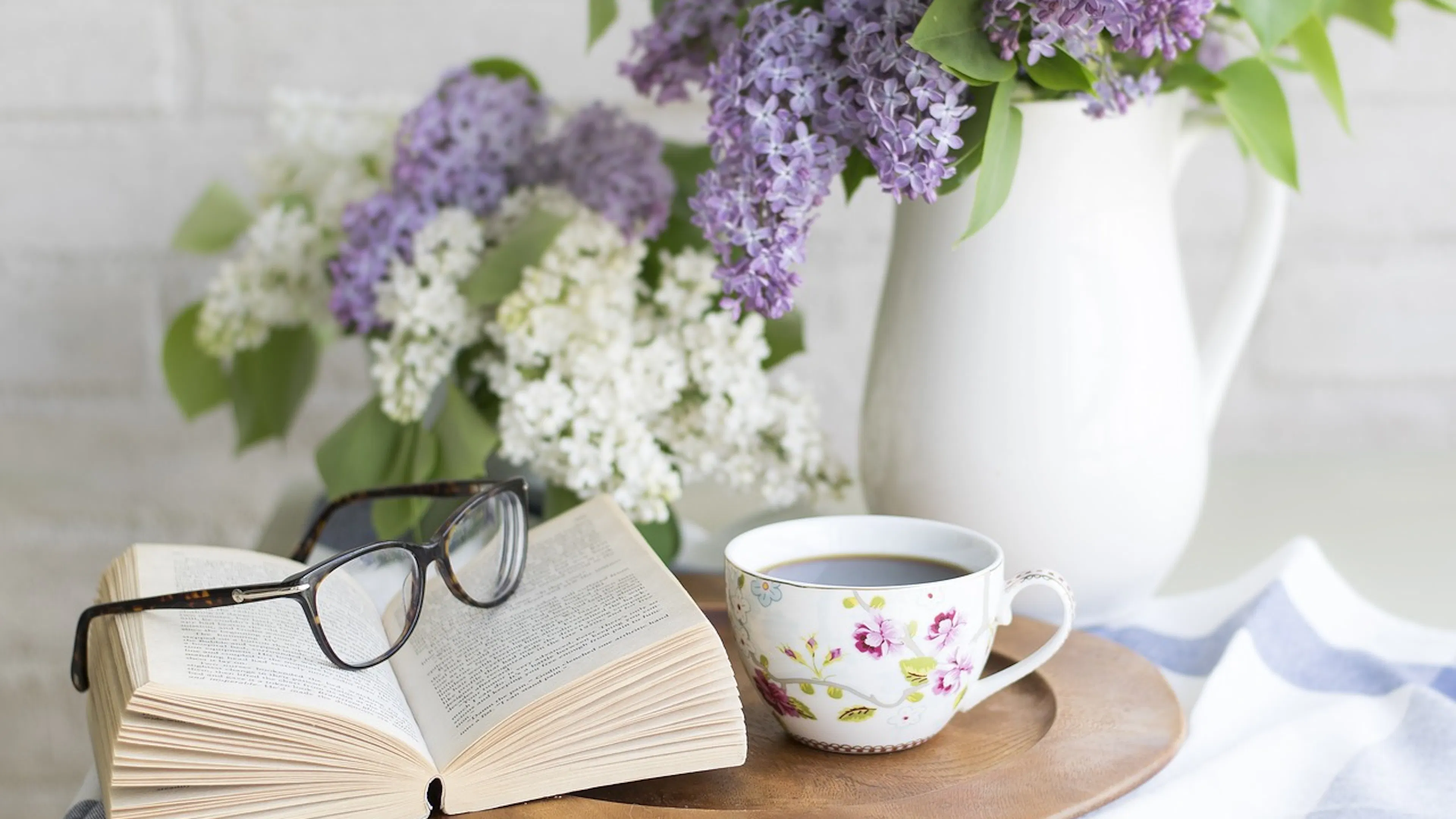 open book with glasses and cup of coffee in flower cup in front of white vase with lilacs
