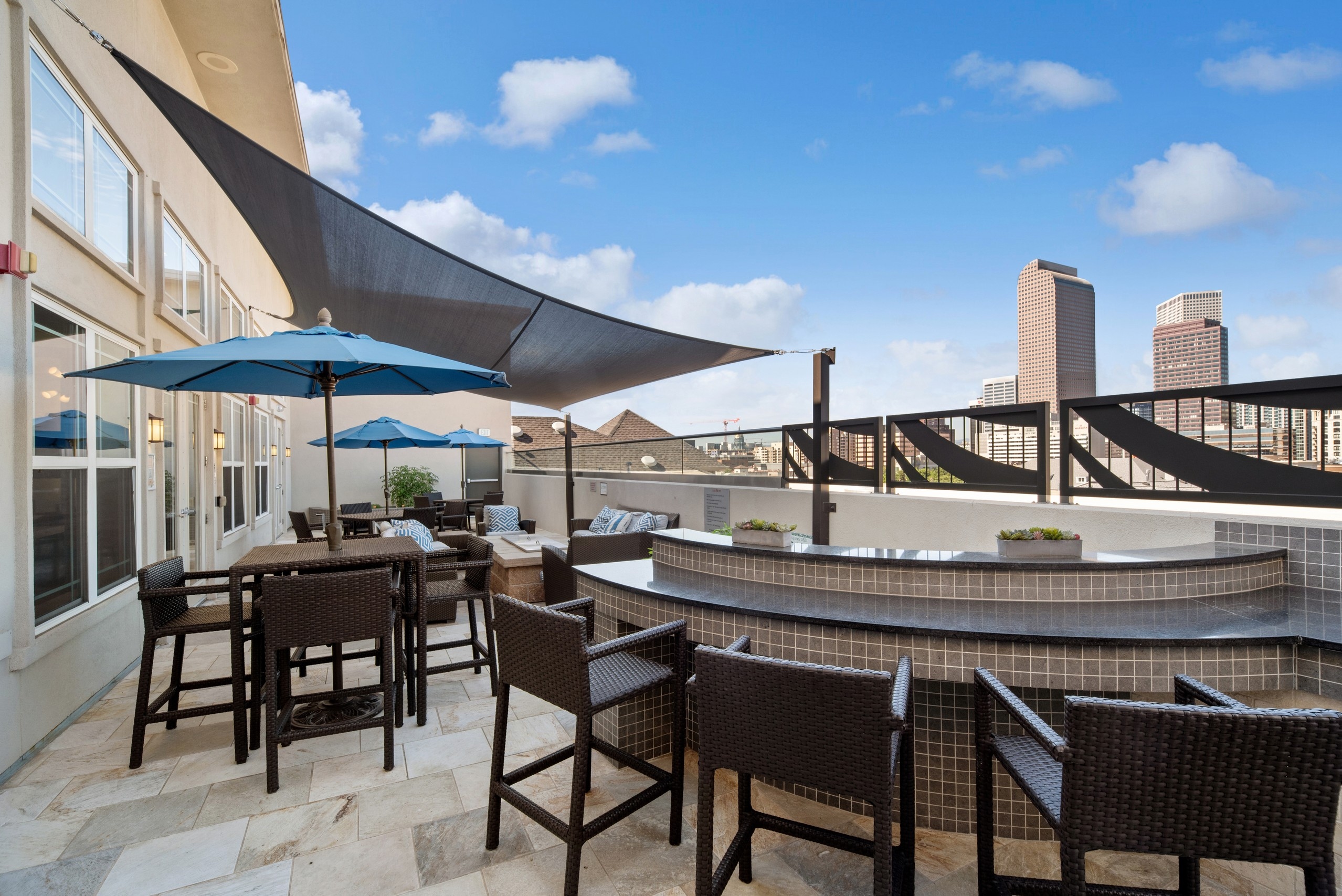Exterior view of a rooftop lounge at AMLI Park Avenue with a bar seating area with chairs and tables with blue umbrellas