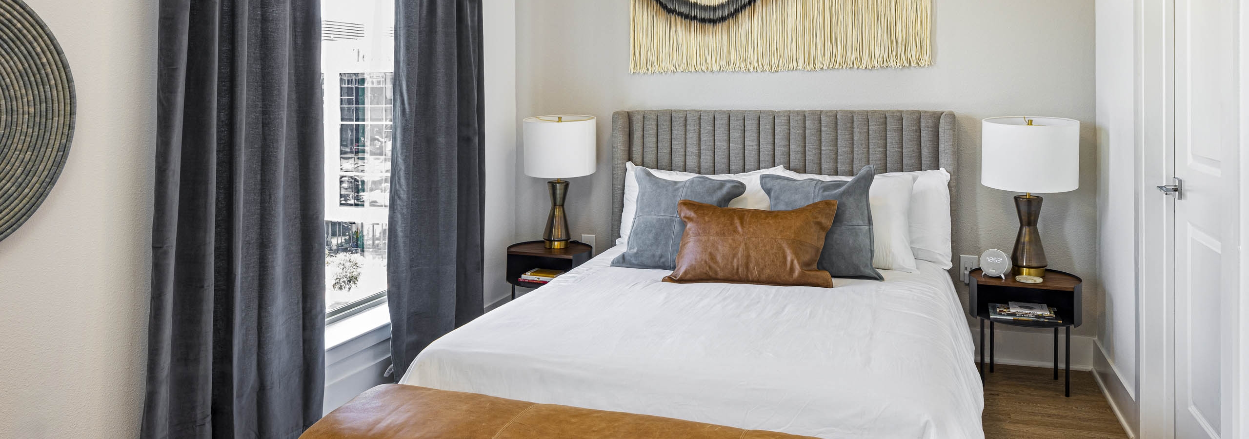 Bedroom with bed and nightstands with lamps and large fringed wall hanging and leather bench at AMLI Branch Park
