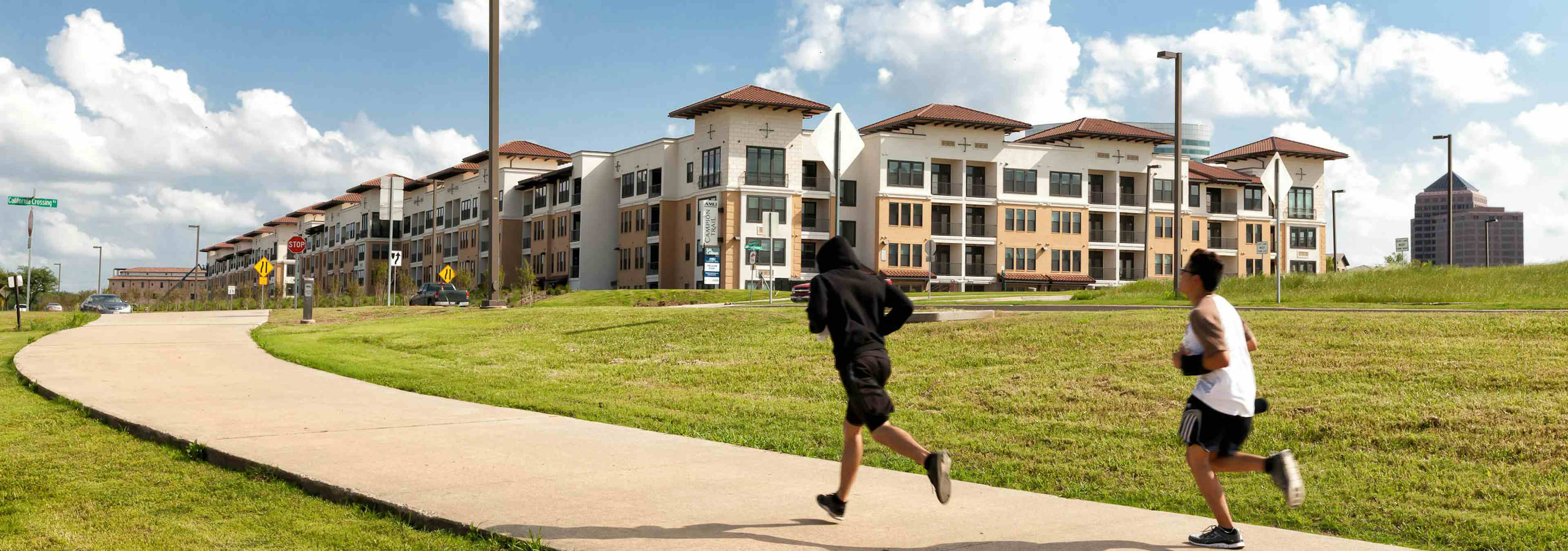 Joggers on concrete path with grass on either side of them and AMLI Campion Trail apartment building in the distance