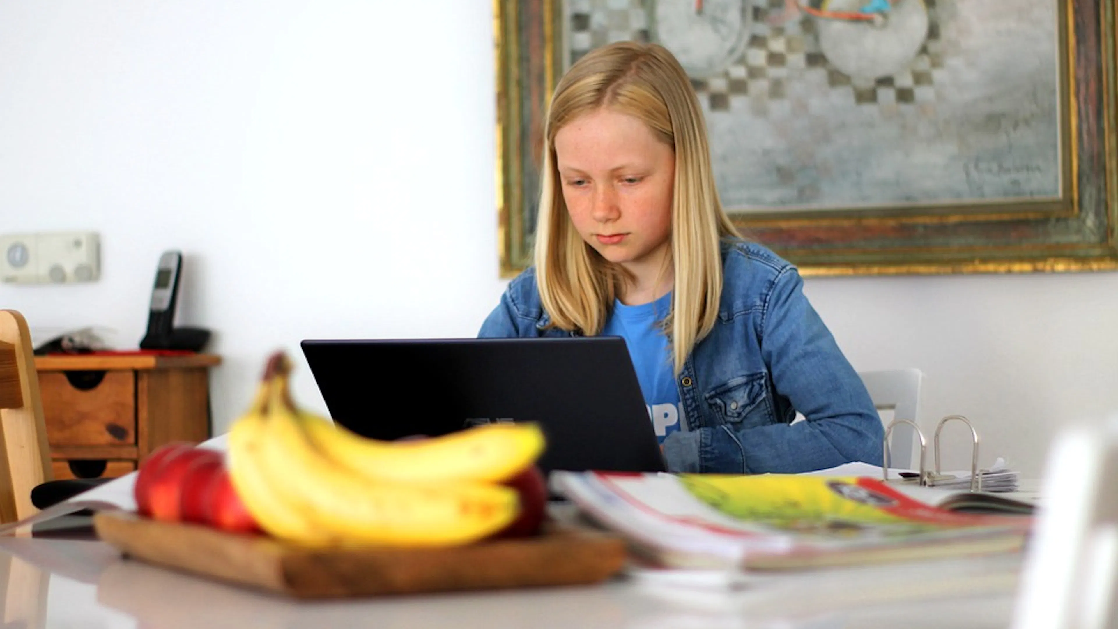A girl sitting at a table in front of a computer with school books and fruit snacks stacked around her as she studies