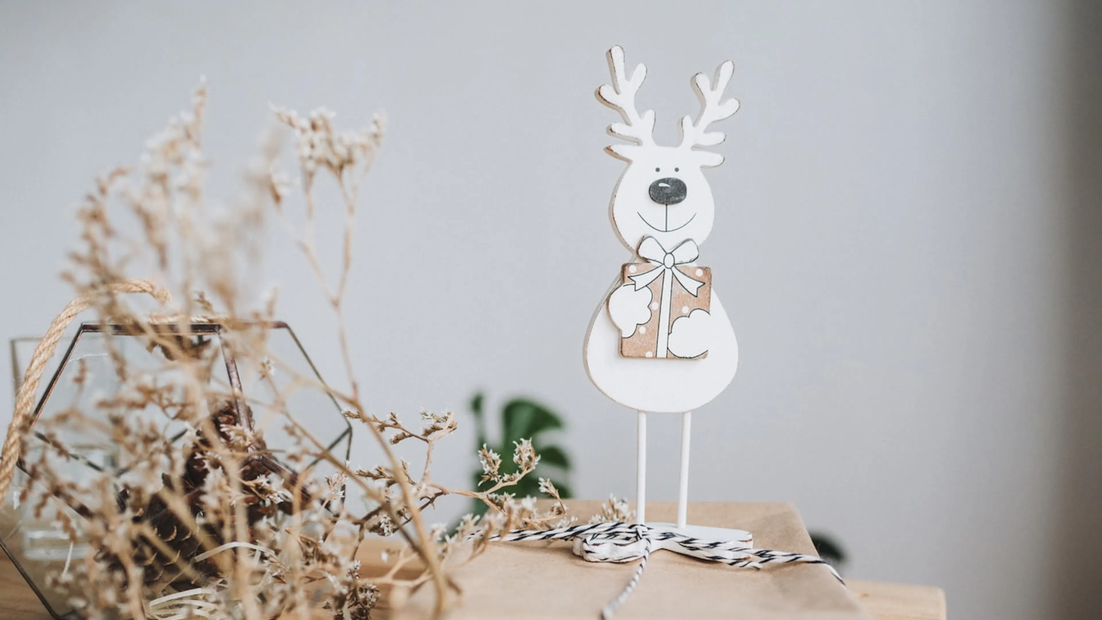 A white wooden reindeer decoration holding a wrapped gift on a wooden table next to a dried bouquet of flowers