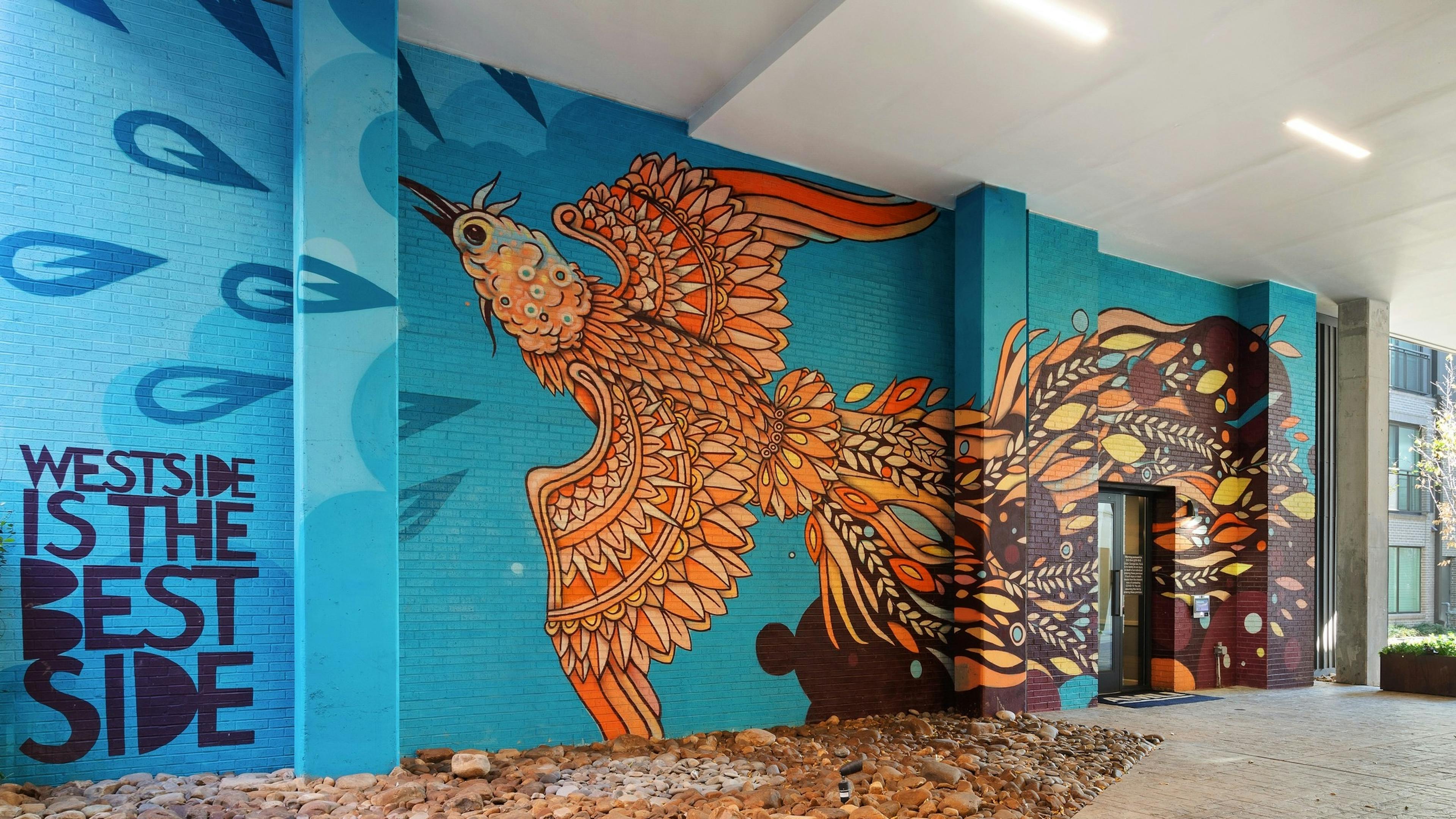 View of bright exterior corridor with beautifully painted mural of flying bird on blue brick wall at AMLI Westside apartments 