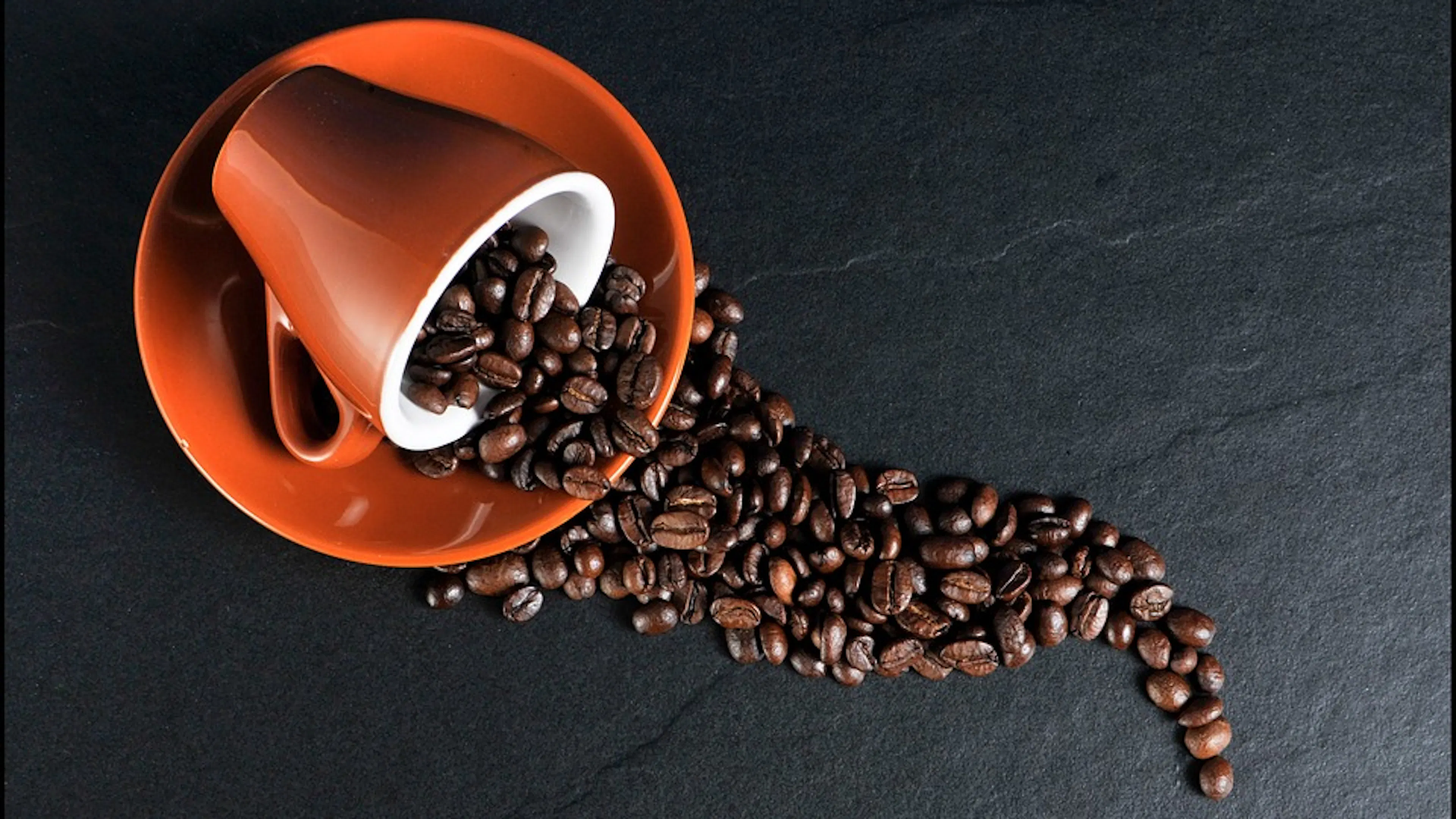 An orange mug and saucer pictured from above with coffee beans spilling out of the overturned cup