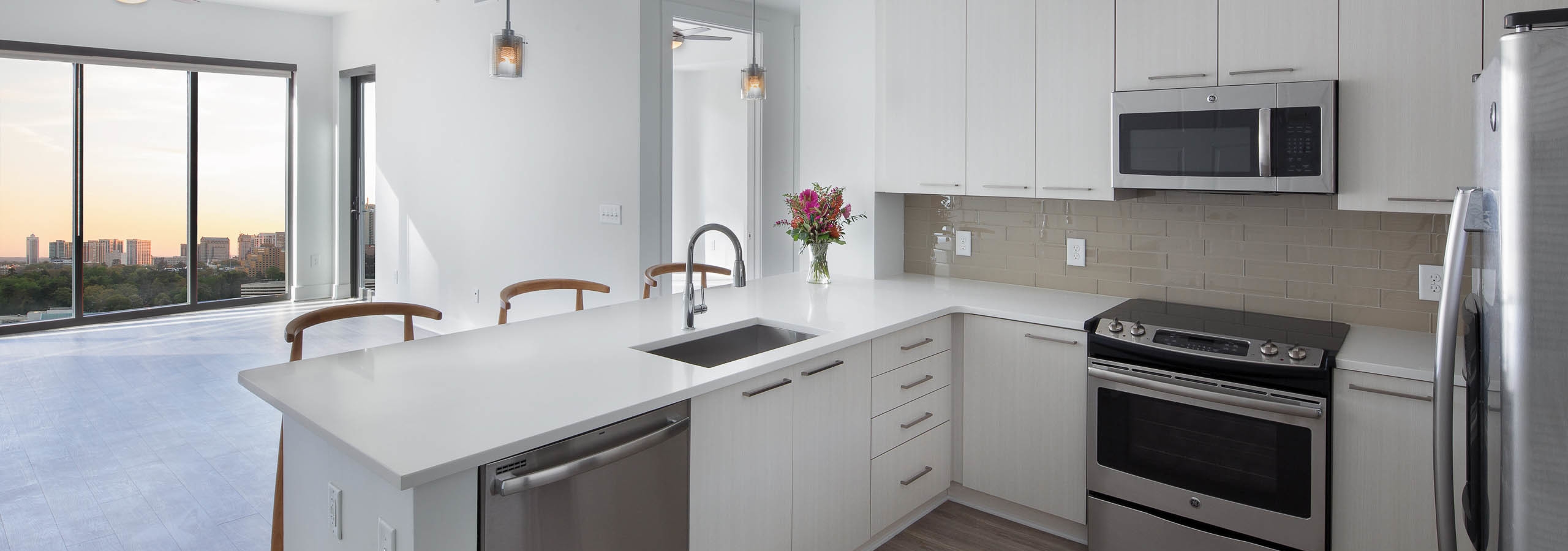 Kitchen at AMLI 3464 with counter and wooden barstools leading into living area with large windows showing a sunset view