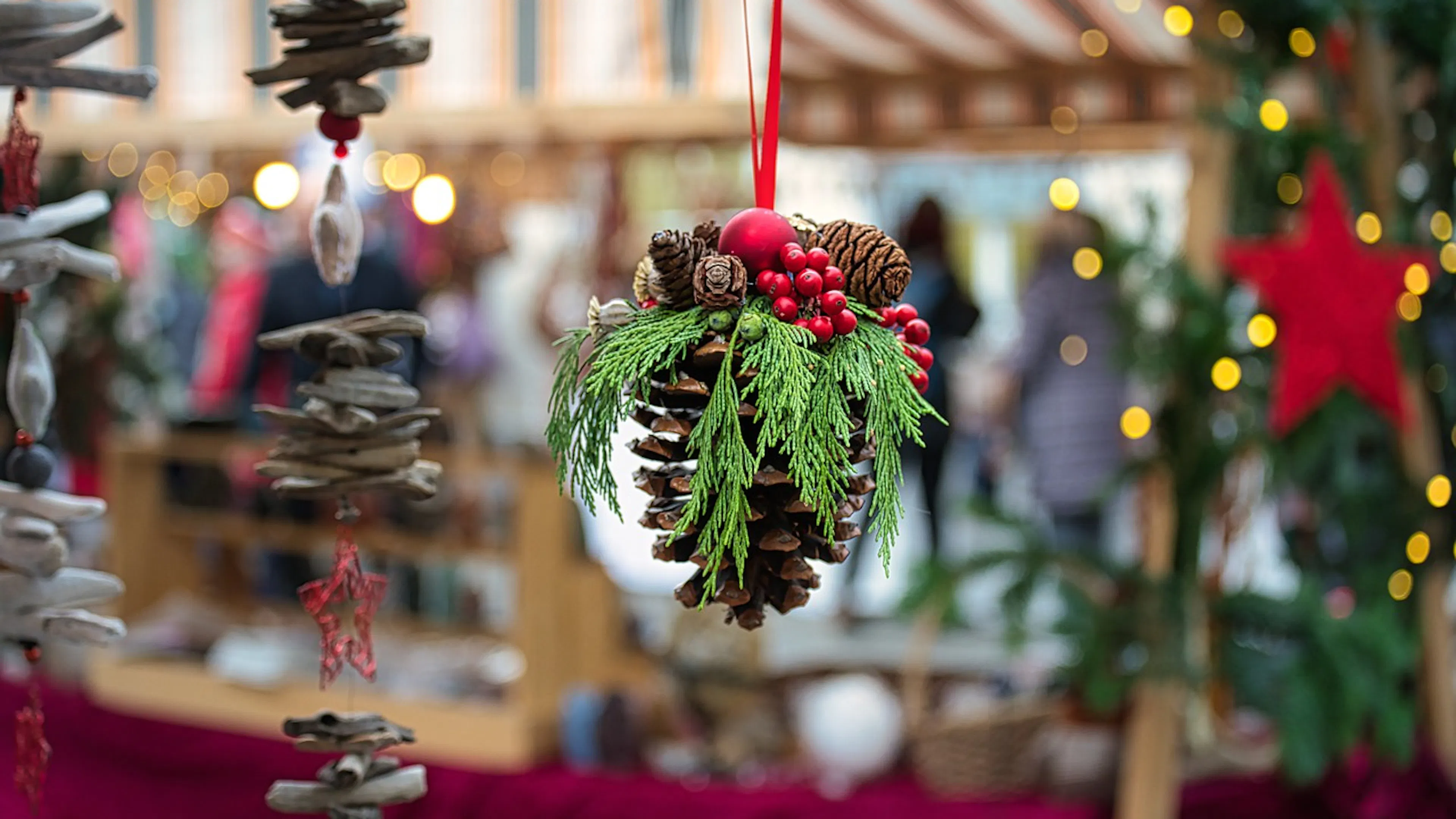 A pinecone hanging from a red ribbon and decorated with cranberries and holly berries in the foreground of a holiday market