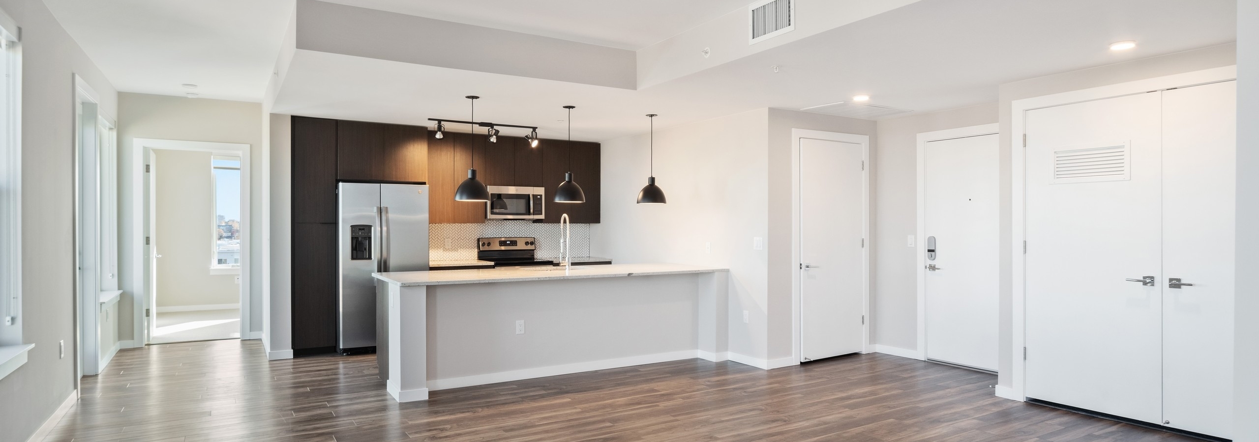 AMLI at Uptown open apartment living area with view of modern kitchen featuring dark cabinetry and modern light fixtures