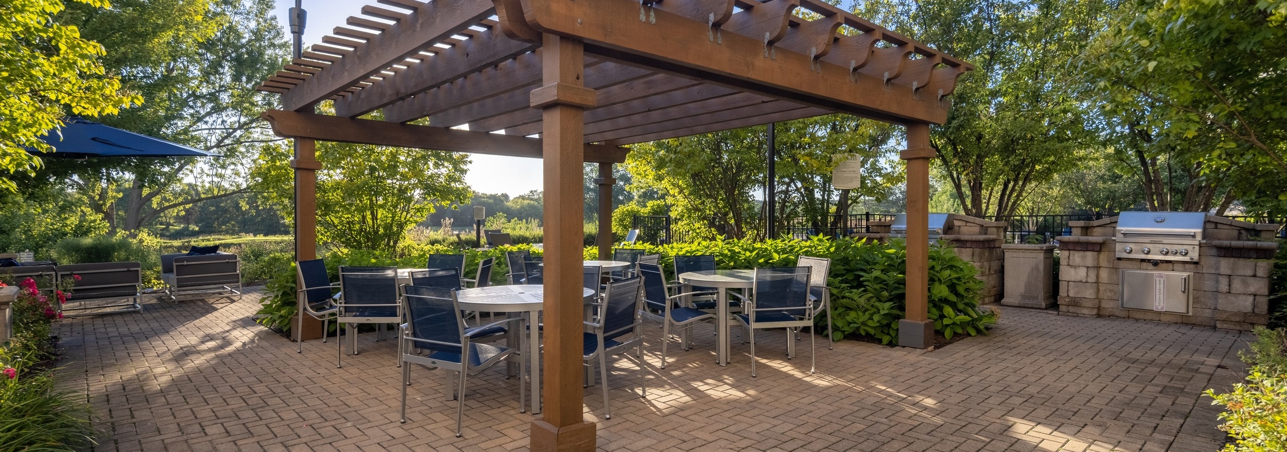 Dual grill station at AMLI at Seven Bridges with tables and hairs under a wood trellis surrounded by green vibrant trees