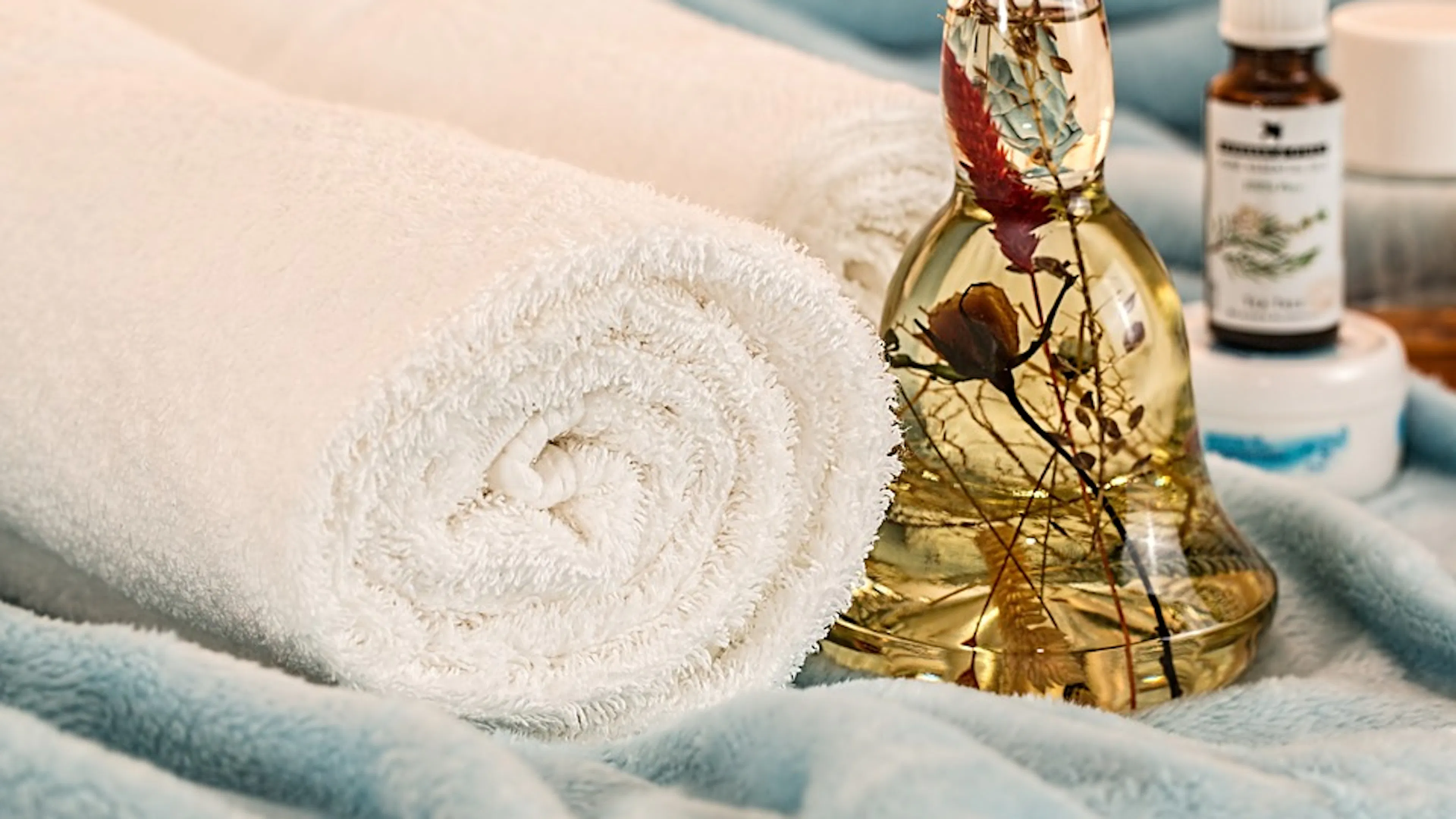Two white towels tightly rolled up and resting on a baby blue towel with a glass jar of floral oil placed next to them