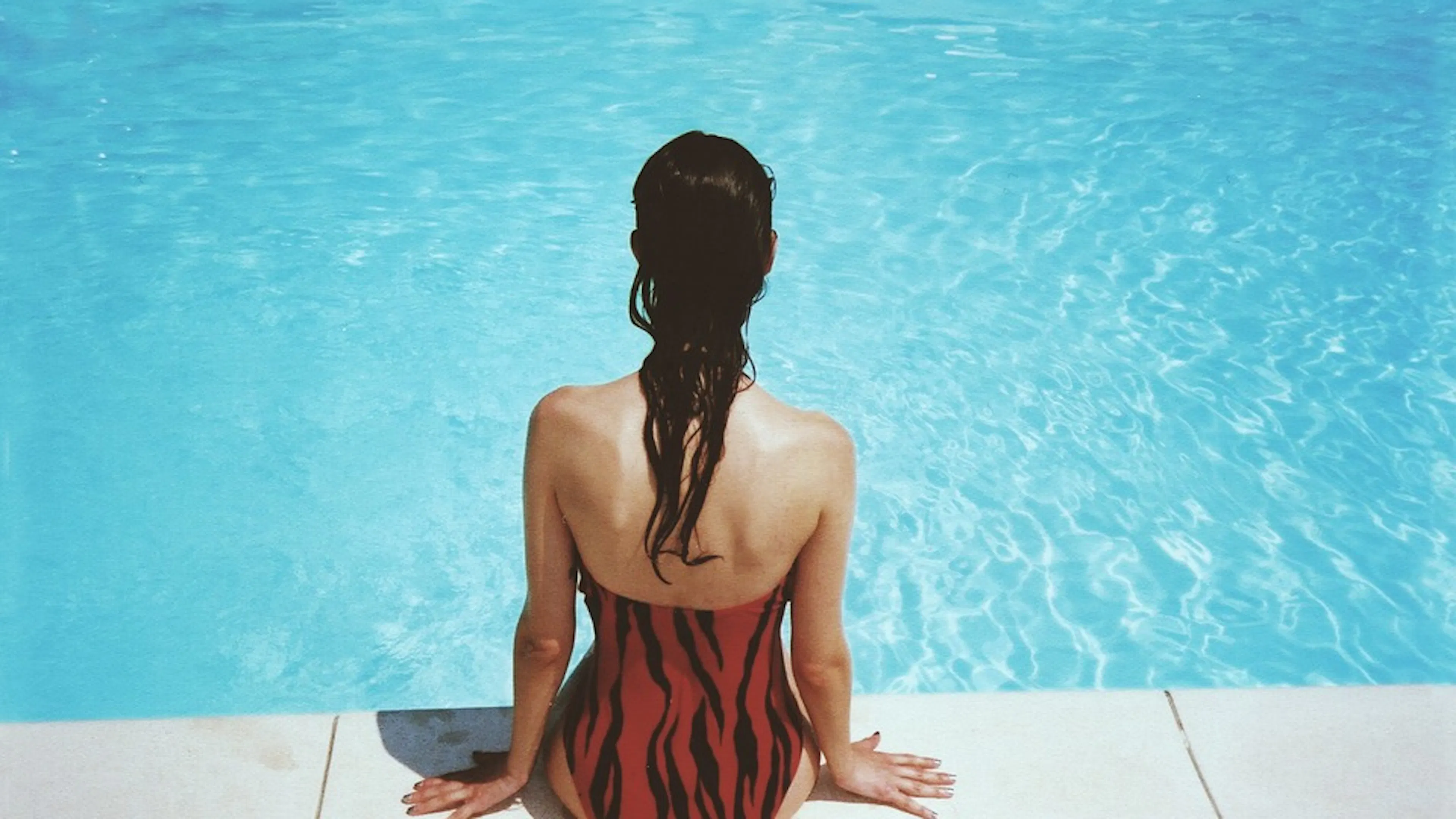 A woman facing away from the camera and sitting at the edge of a swimming pool