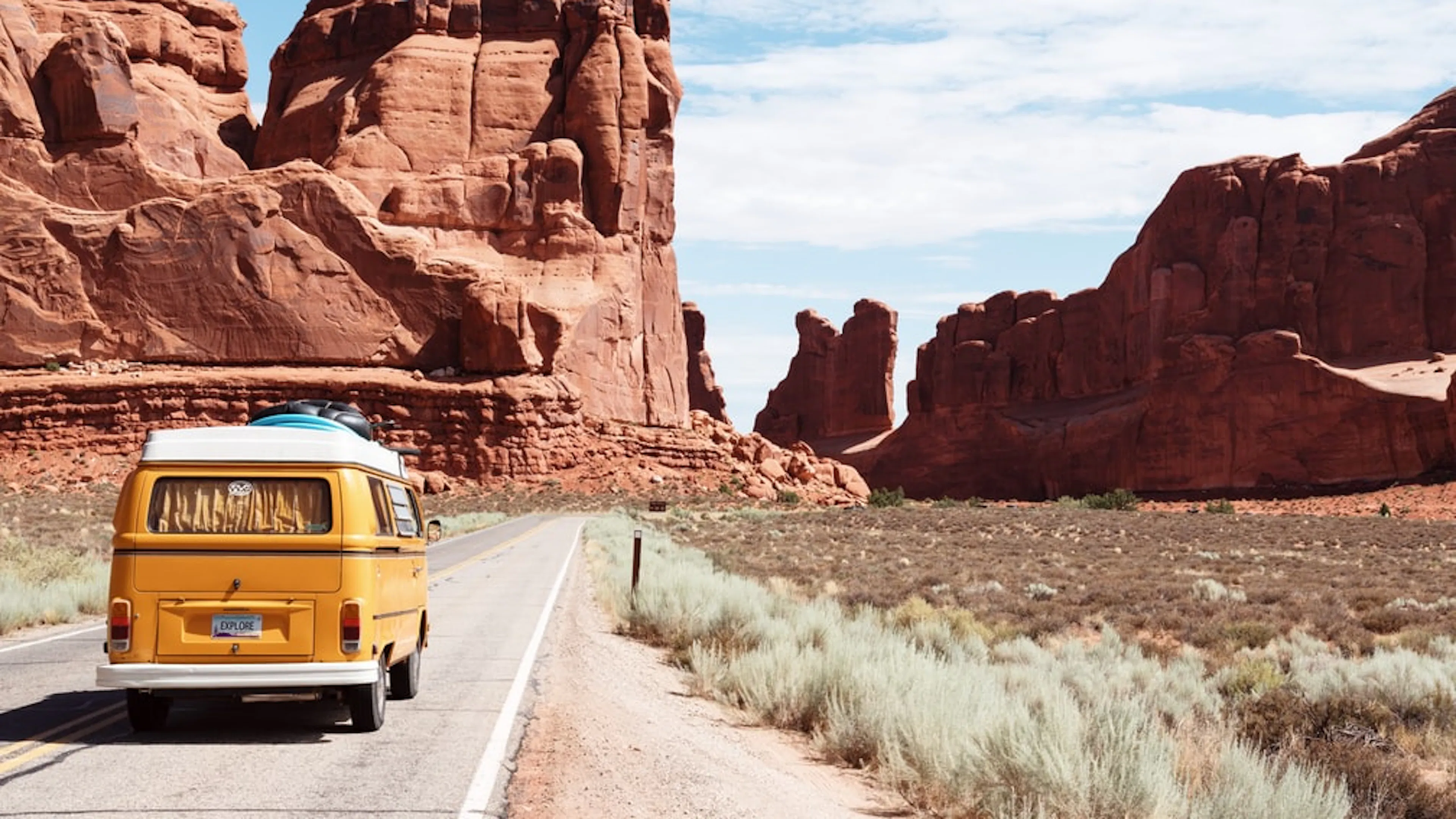 An old yellow camper van driving away toward some barren desert cliffs surrounded by low dry shrubs and red sand